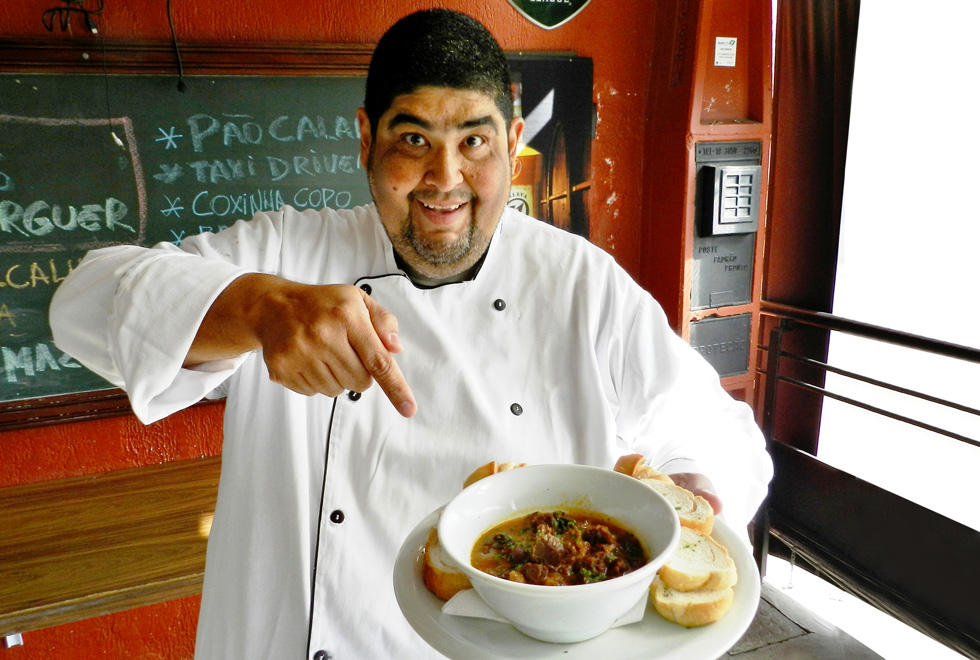 Manuel Alves, vestido com as roupas de chef, sorri e aponta para um prato 