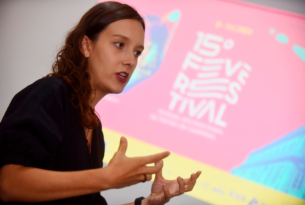 audiodescrição: fotografia colorida de bruna schroeder, que gesticula e está falando. ao fundo, uma projeção do cartaz do festival internacional de teatro de campinas, nas cores rosa, azul e amarelo.