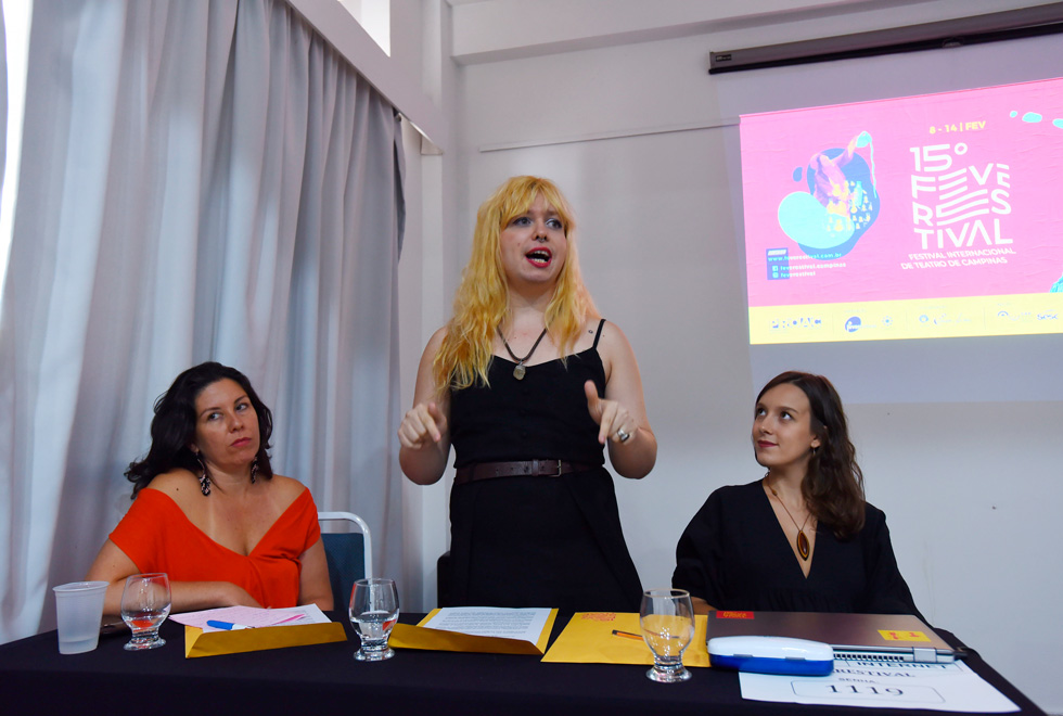 audiodescrição: fotografia colorida mostra uma mesa com três mulheres. a do meio está em pé, gesticulado e olhando para a frente. ela é helena agalanéa, uma das curadoras do 15º festival internacional de teatro de campinas.