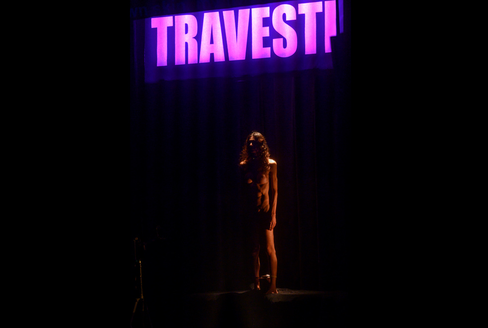 audiodescrição: fotografia colorida de cena do espetáculo Manifesto transpofágico escura, com fundo preto. há um letreiro luminoso, cor lilás neon, escrito travesti. uma pessoa nua, de cabelos longos e semi-iluminada, está abaixo do letreiro.