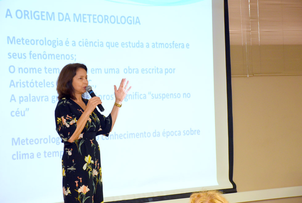 Ana Ávila em frente ao telão iluminado por um slide com informações sobre a origem da meteorologia