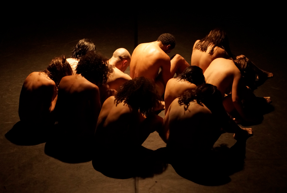 Dez bailarinos estão sentados no chão. A luz do espetáculo ilumina mais as costas de alguns deles.