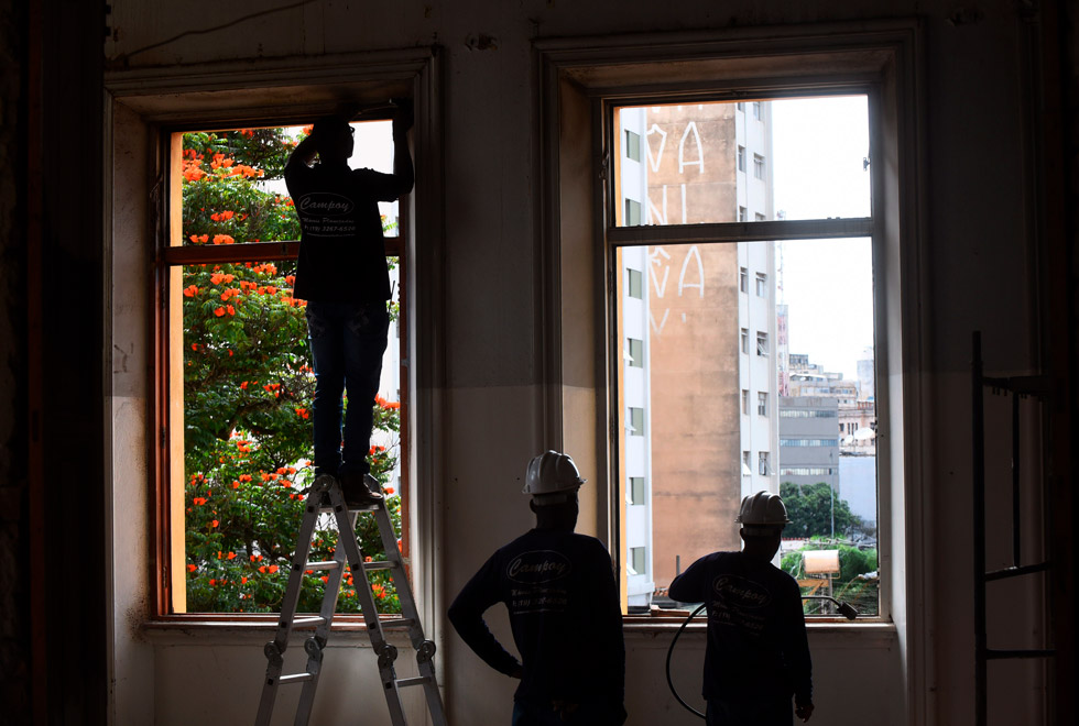 audiodescrição: fotografia colorida mostra trabalhadores de costas em obras na janela de uma casa