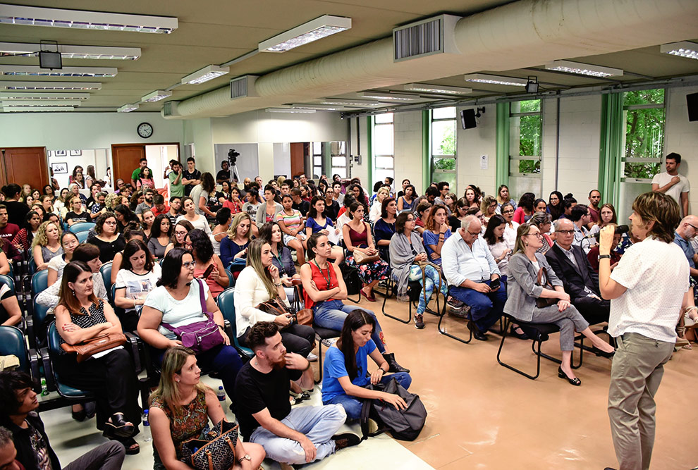 Foto mostra auditório cheio com pessoas inclusive sentadas no chão e, à frente, fala ao microfone a coordenadora do evento