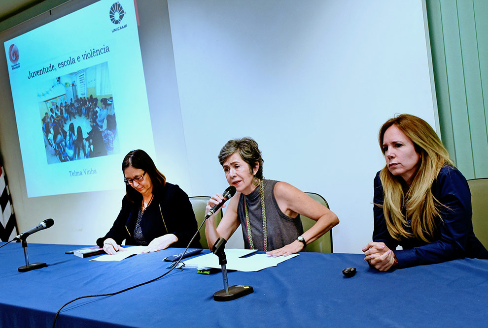 Imagem mostra as três participantes da primeira mesa redonda sentadas. Ao fundo está o telão onde está escrito Juventude, escola e violência