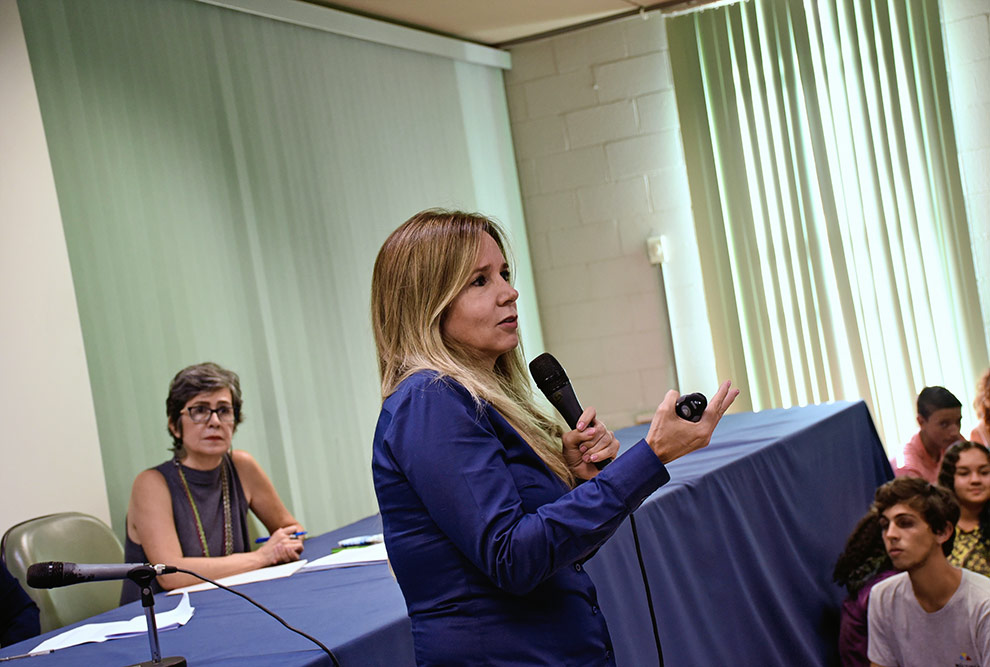 Telma Vinha, com o microfone, fala ao publico. Ela ocupa o centro da imagem. à esquerda uma professora que estava na mesa de abertura a observa. Há também pessoas sentadas no chão à direita.