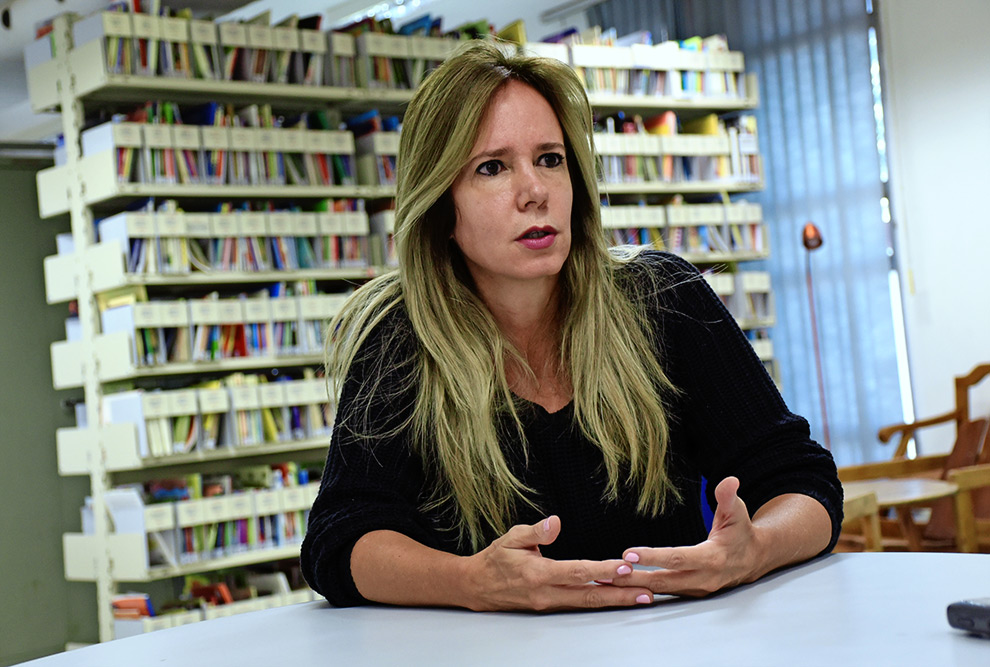 Telma Vinha durante a entrevista. Ela é loira cabelos lisos, está maquiada e séria durante entrevista na biblioteca da Faculdade de Educação