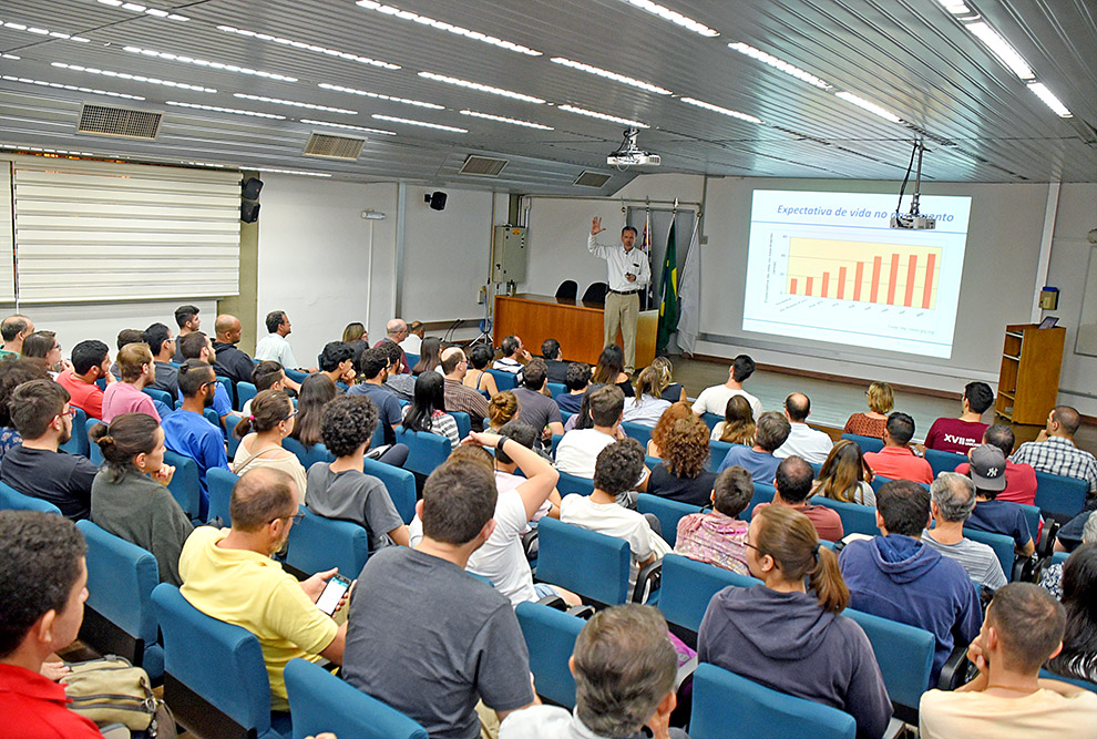 Plateia no auditório assiste a palestra de Brito Cruz. A foto mostra várias pessoas de costas, sentadas, e à frente o palestrante, em frente ao telão, levantando uma das mãos