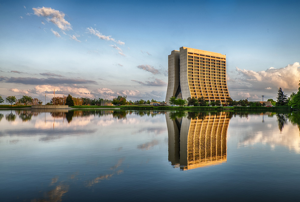 Foto de divulgação mostra edifício do Fermilab nos Estados Unidos ao fundo. O prédio tem arquitetura não convencional com a base mais alargada. O prédio tem a imagem refletida em um grande lago.