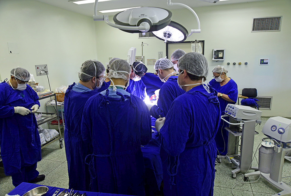 Equipe do centro cirúrgico do Hospital de Clínicas da Unicamp realiza cirurgia. Na foto, nove profissionais estão paramentados com jalecos azuis e toucas brancas em volta de um paciente que está sendo operado na mesa.