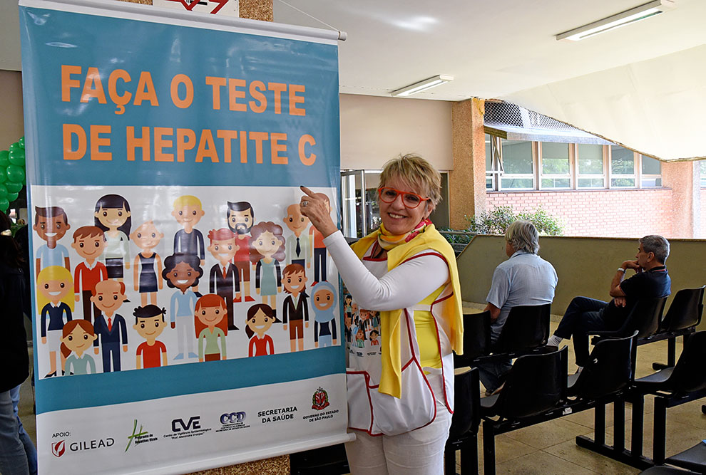 Rachel Stucchi, em pé mostra o cartaz da campanha onde está escrito "faça o teste da hepatite C"