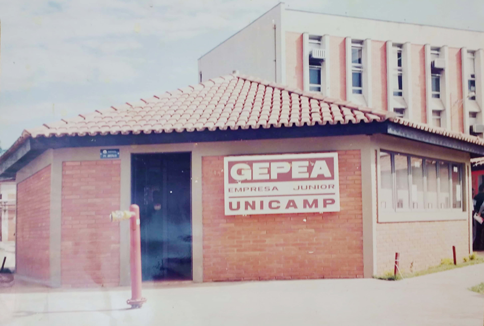 Imagem mostra prédio antigo da Gepea, primeira empresa júnior da Unicamp fundada em 1990 na Faculdade de Engenharia de Alimentos. Há uma porta ao centro da sede. À esquerda consta uma placa patrimonial do imóvel. À direita vem a inscrição Gepea - Empresa Junior - Unicamp. Ao fundo aparece parte de um dos pre´dios da Faculdade de Enegenharia de Alimentos da Unicamp. AQmbas as construções possuem, acabamento com tijolinhos à vista