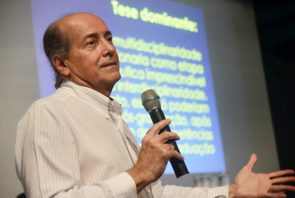 Naomar de Lameida durante sua palestra, Na foto ele está à esquerda, segurando um microfone e com braços abertos, com a tela onde eram apresentados os slides ao fundo, 