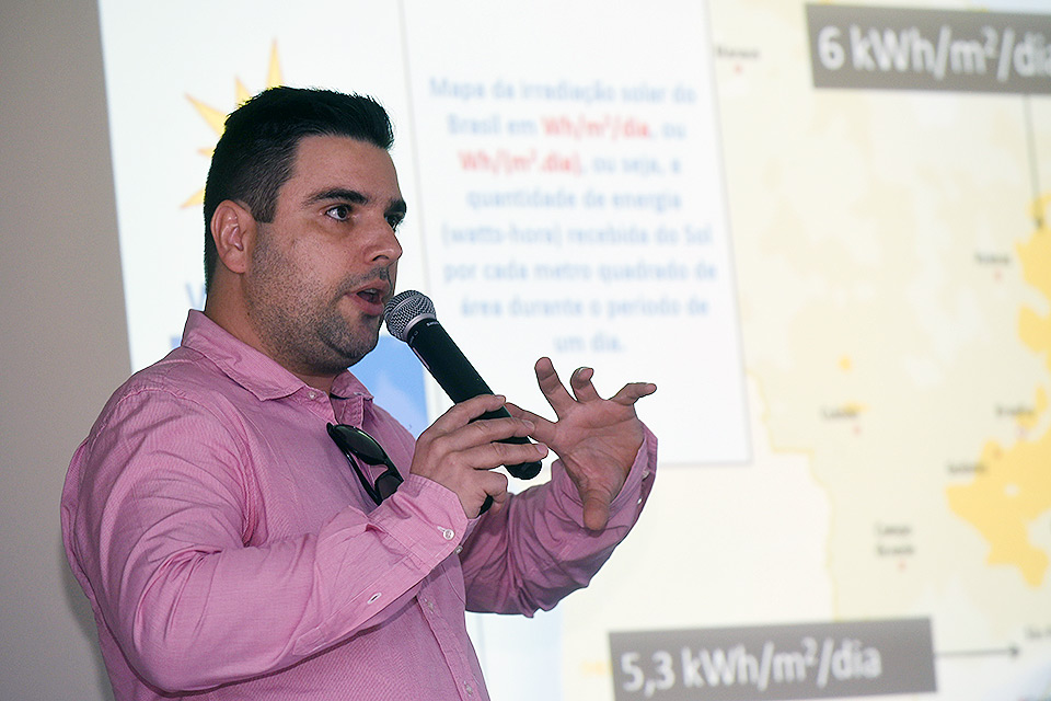 Marcelo Villalva gesticula em frente ao painel iluminado na palestra que deu na Educorp. Ele veste uma camisa de botão cor de rosa e tem um oculos de sol pendurado no colarinho