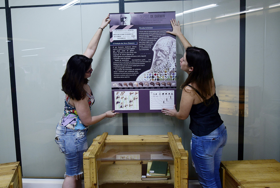 Duas mulheres colocam na parede um dos painéis da exposição