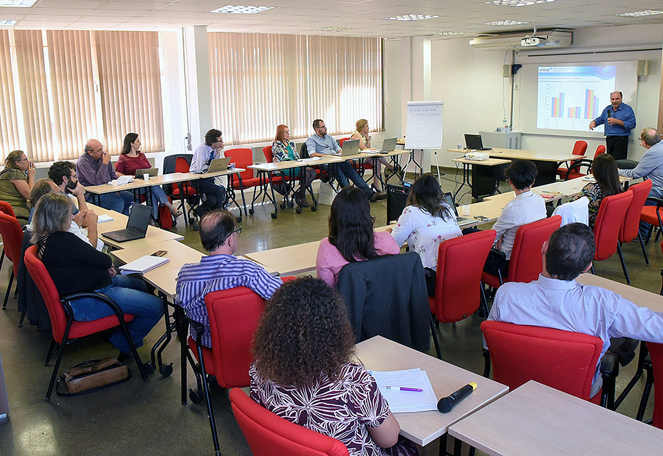 Participantes da reunião estão sentados assistindo a apresentação do representante da Unesp