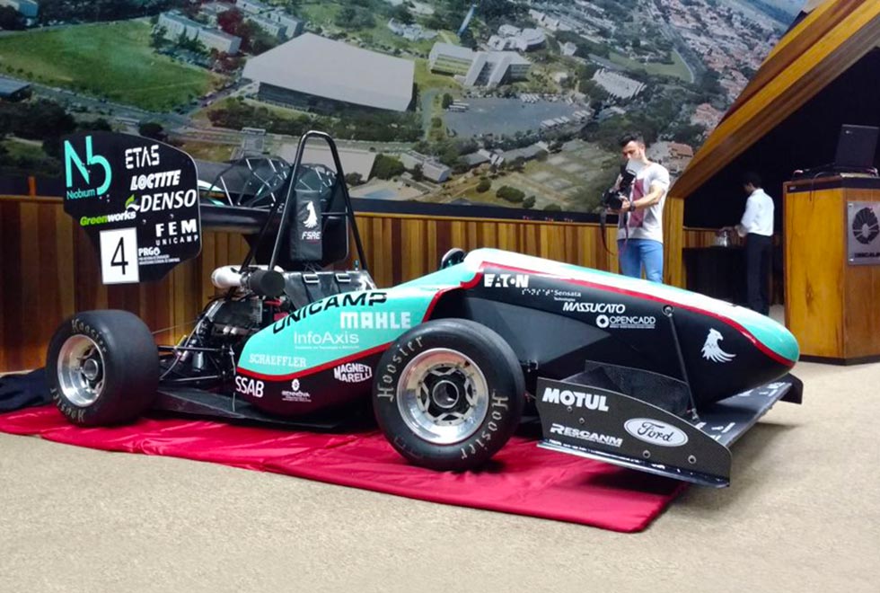 Imagem do carro protótipo F2017, construído pela equipe de estudantes FSAE Unicamp. Na foto o carro monoposto está de lado, voltado para a direita, em cima do palco do auditório do centro de convenções sobre um tapete vermelho. O carro é cinza e preto, com cockpit aberto e em sua carenagem consta os nomes das empresas patrocinadoras