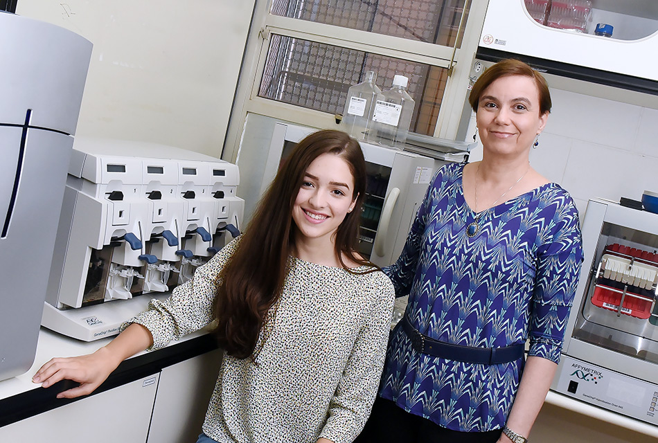 A aluna Caroline está sentada tendo ao lado a professora Iscia. Imagem feita no Laboratório. Ao fundo estão equipamentos na bancada.