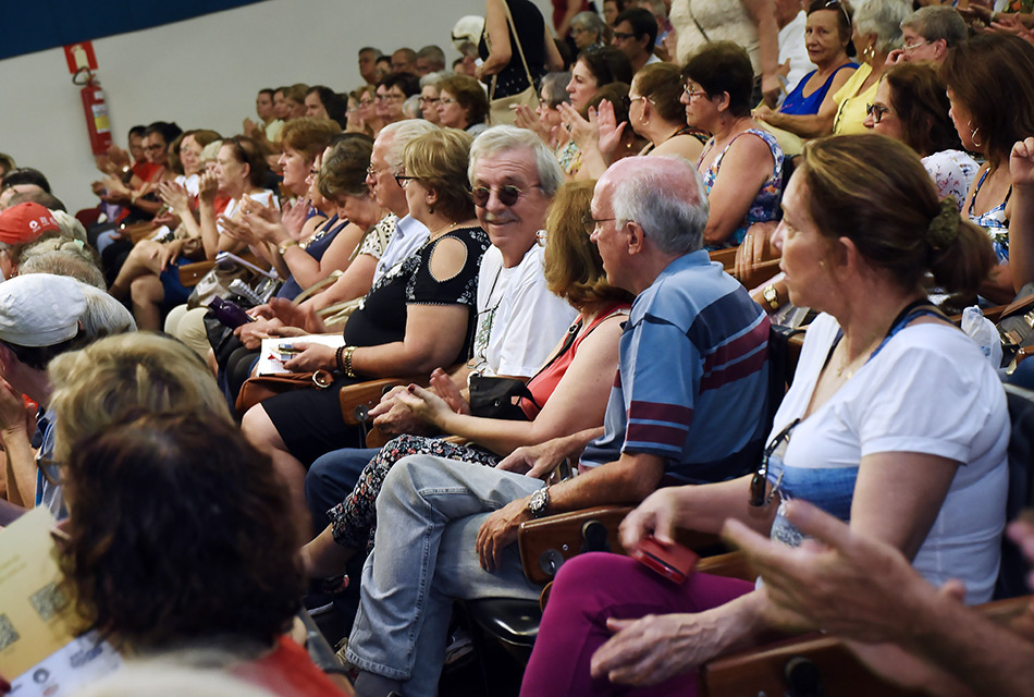 senhoras e senhores de idade assistem palestra em auditório sentados. Alguns riem, outros batem palmas outros conversam.