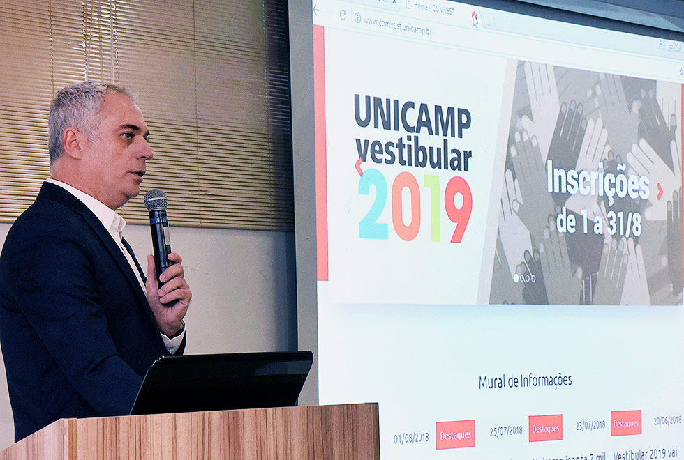 José Alves de Freitas Neto segura o microfone e fala à plateia da DGA. Ao fundo a imagem no telão é de um slide onde está escrito "Vestibular Unicamp 2019, Inscrições de 01 a 31 de agosto