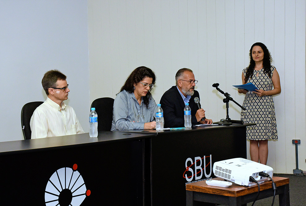 Mesa de abertura do evento com três pessoas sentadas e a docente coordenadora em pé ao lado direito 