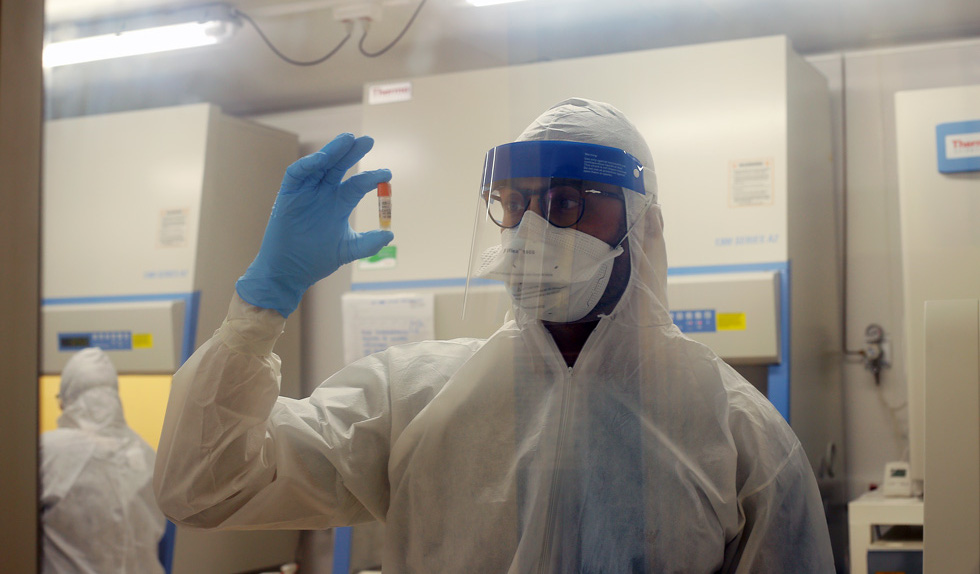 foto mostra pesquisador vestido com roupas de proteção segurando uma amostra de coronavírus