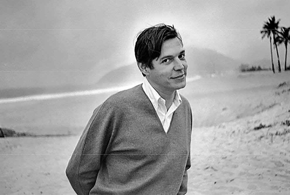 Tom Jobin, fotografado em preto e branco, da cintura para cima, em uma praia. Ele é branco, tem o cabelo curto, usa blusa de manga comprida e está sorrindo.