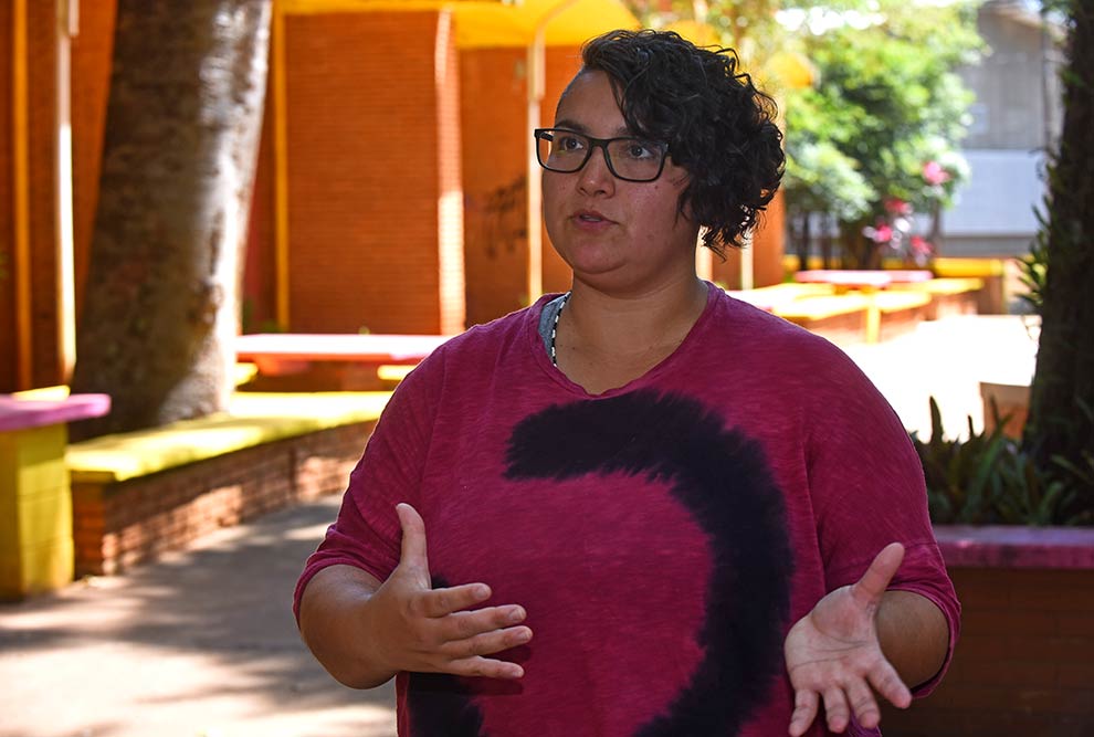 Foto de uma mulher que aparece da cintura para cima. Ela é branca, tem o cabelo curto e preto. Está usando óculos e uma blusa pink.