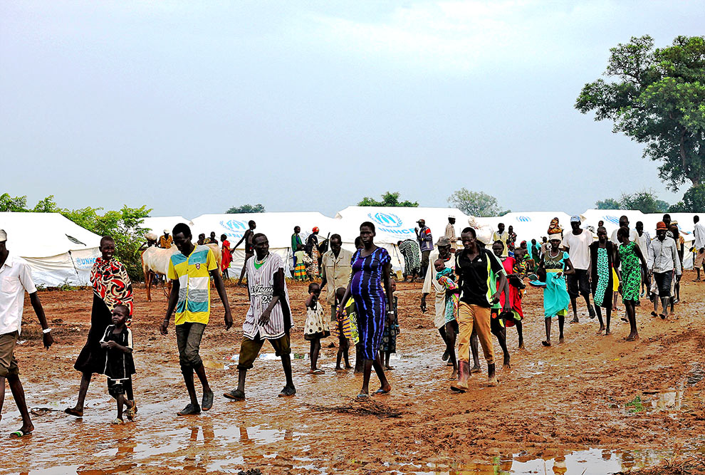 foto mostra pessoas refugiadas andando em um campo aberto com tendas da onu ao fundo