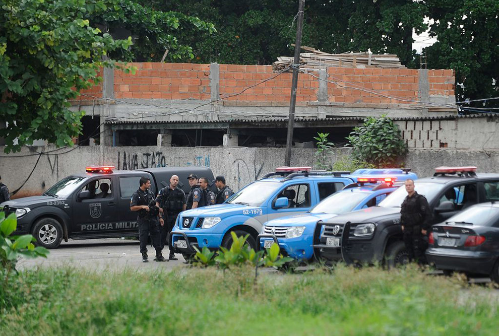 Foto que mostra carros de polícia estacionados em uma rua. Há policiais em pé ao lado dos carros. Ao fundo há um muro e uma casa com paredes sem reboco.