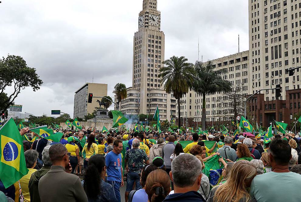 Foto de um manifestantes em frente a um prédio do governo. Eles usam camisas nas cores verde e amarelo e seguram bandeiras do Brasil.