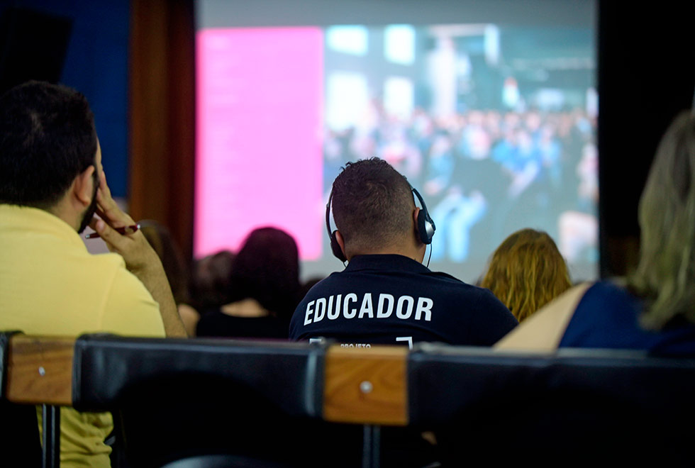 Foto mostra rapaz de costas na plateia. Em sua camiseta está escrita a palavra educador