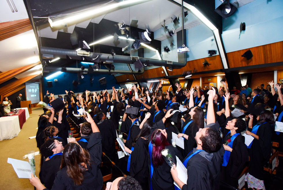 audiodescrição: fotografia colorida de diversos estudantes de toga em auditório comemorando formatura, jogando os chapeus para cima