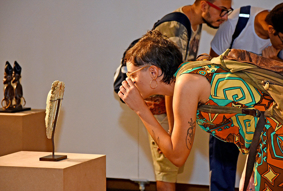 audiodescrição: fotografia colorida mostrando jovem olhando para uma escultura em um local de exposição artística