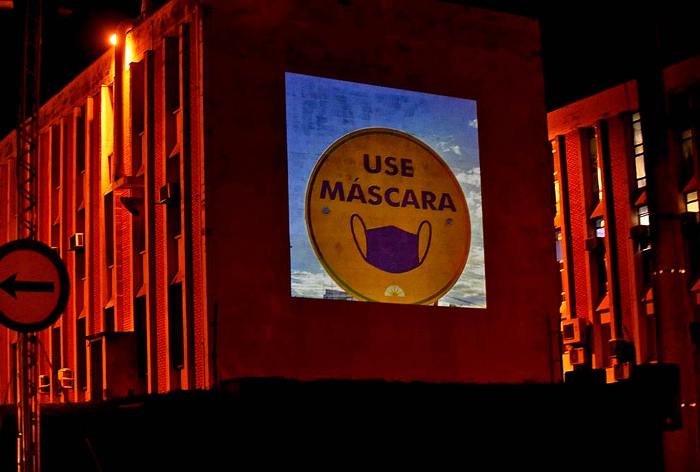 foto mostra ambiente noturno, com uma projeção realizada em um prédio com imagem "use máscara"