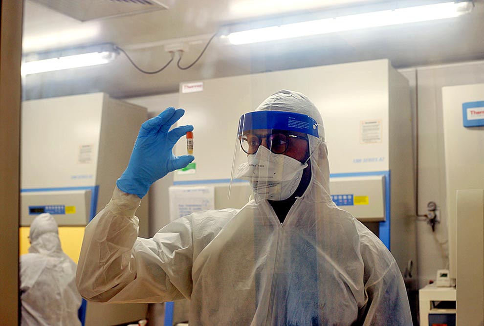 foto mostra pesquisador vestido com roupas de proteção segurando uma amostra de coronavírus