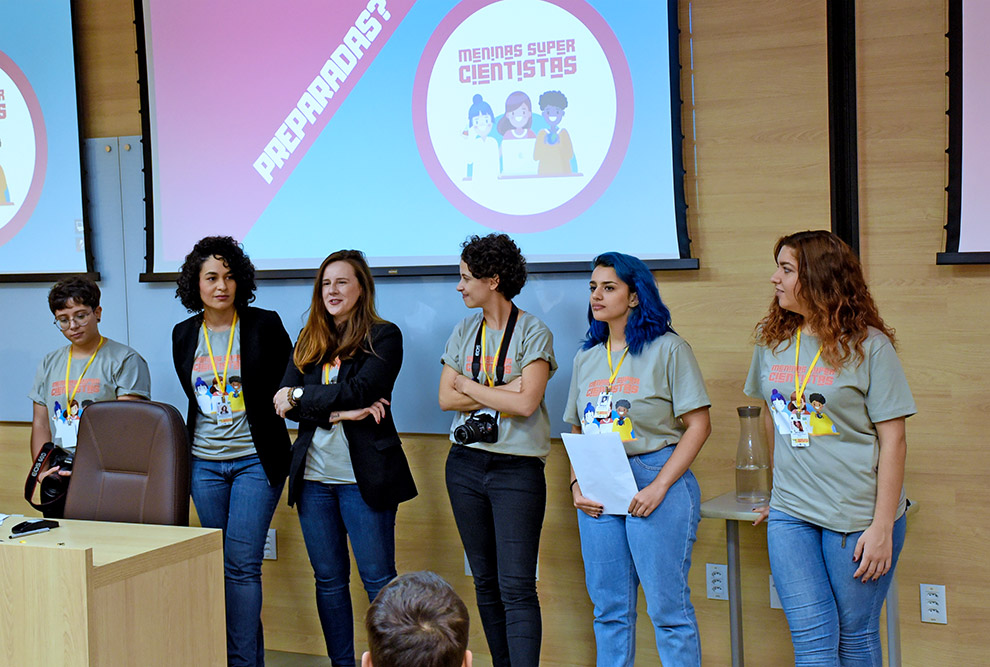 audiodescrição: fotografia colorida mostra seis mulheres em pé em cima de um palco. elas são as organizadoras do meninas super cientistas