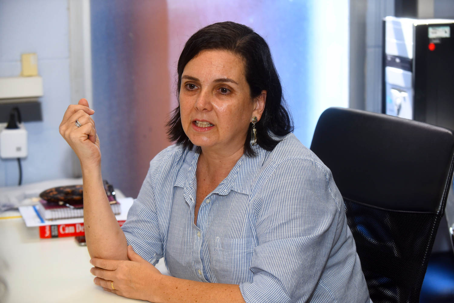 audiodescrição: fotografia colorida mostra professora Kátia Lucchesi, sentada à mesa, enquanto dá entrevista.