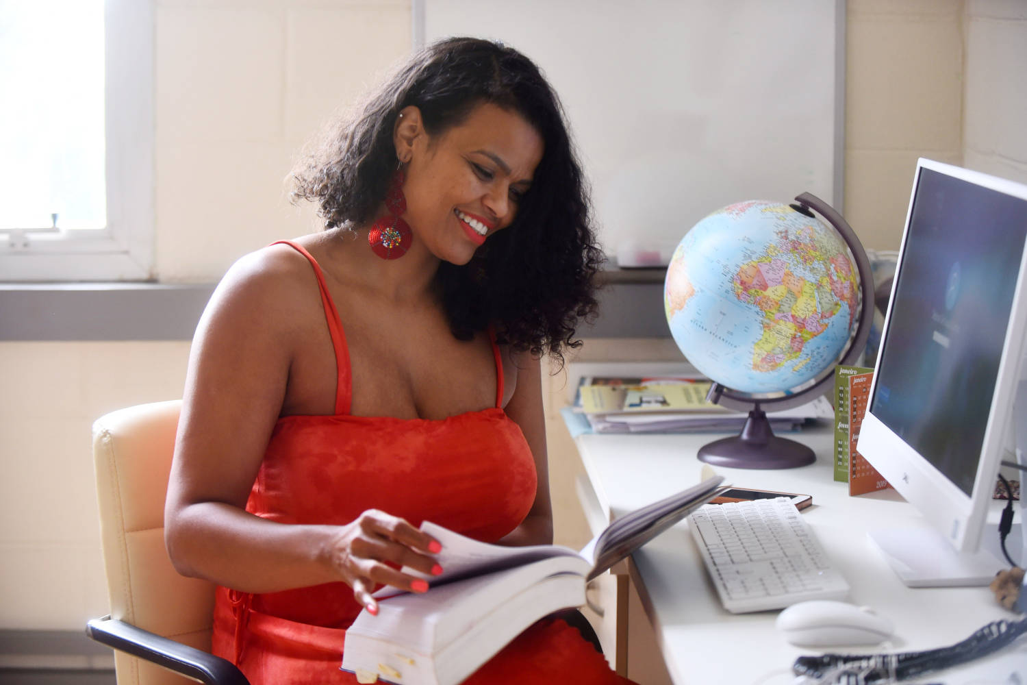 audiodescrição: fotografia colorida mostra professora Débora folheando livro sentada em uma mesa em frente ao computador