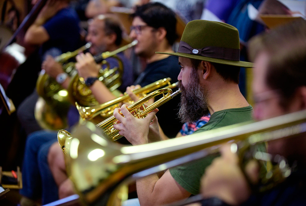 imagem mostra músicos tocando instrumentos de sopro. Um deles está focado, mais ao centro, e usa um chapéu. Os outros estão um pouco desfocados