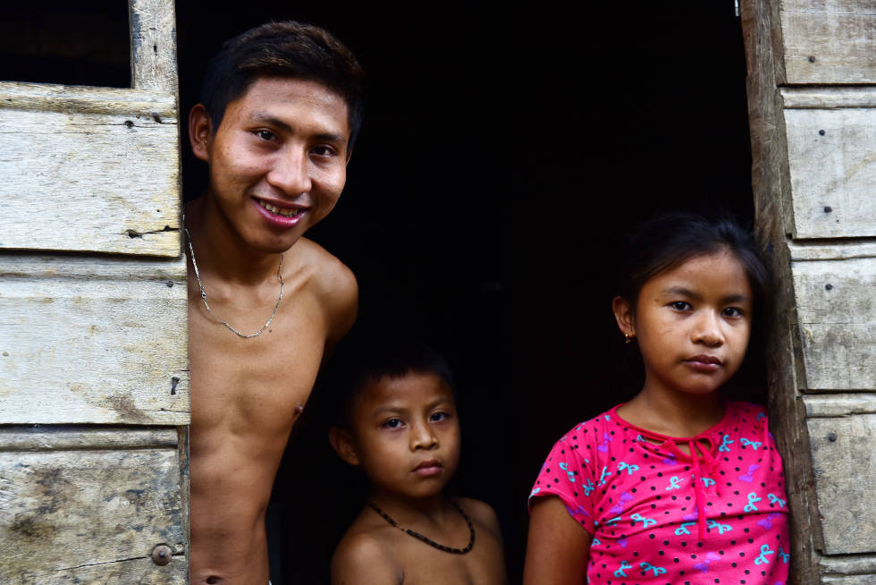 audiodescrição: fotografia colorida de três crianças indígenas posando para a foto em frente a uma casa de madeira