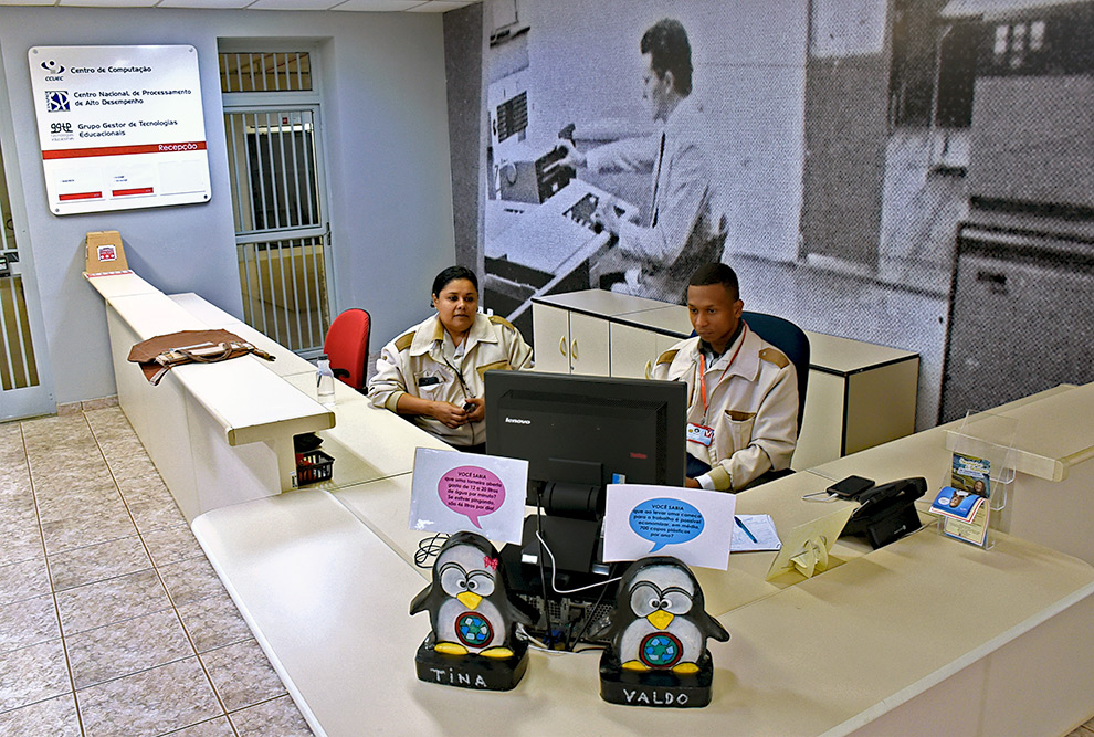 Esculturas de dois Pinguins com os nomes Valdo e Tina estão sobre o balcão da recepção do CCUEC. Atrás do balcão estão dois recepcionistas, um homem e uma mulher