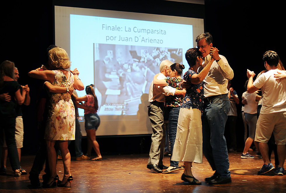 Vários casais dançam tango n palco do auditório do Instituto de Artes, ao fundo o telão exibe um slide da apresentação onde está escrito Finale: La Cunparsita por Juan D'Arienzo