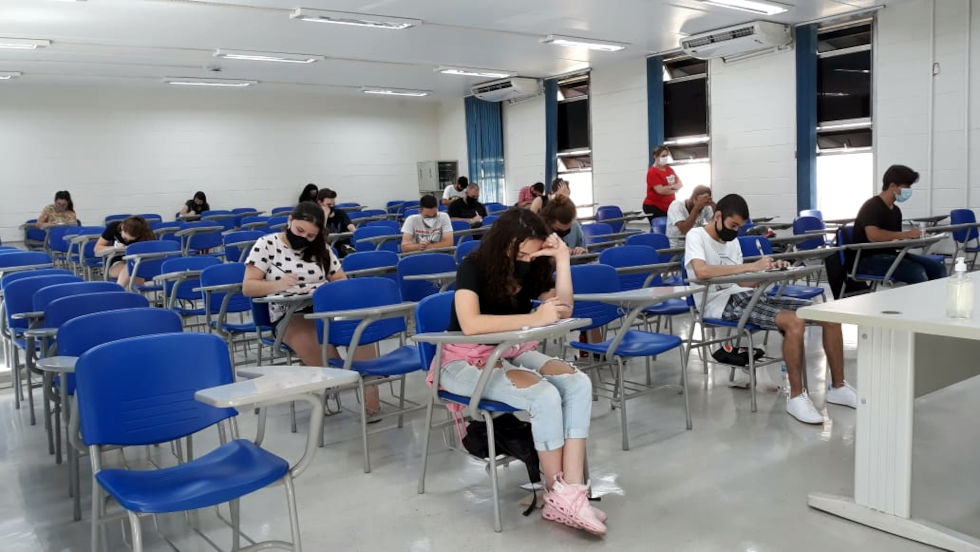 audiodescrição: fotografia colorida mostra estudantes fazendo prova de vestibular; eles estão de máscaras, as carteiras são azuis e há poucos candidatos na sala, conforme protocolos de distanciamento social