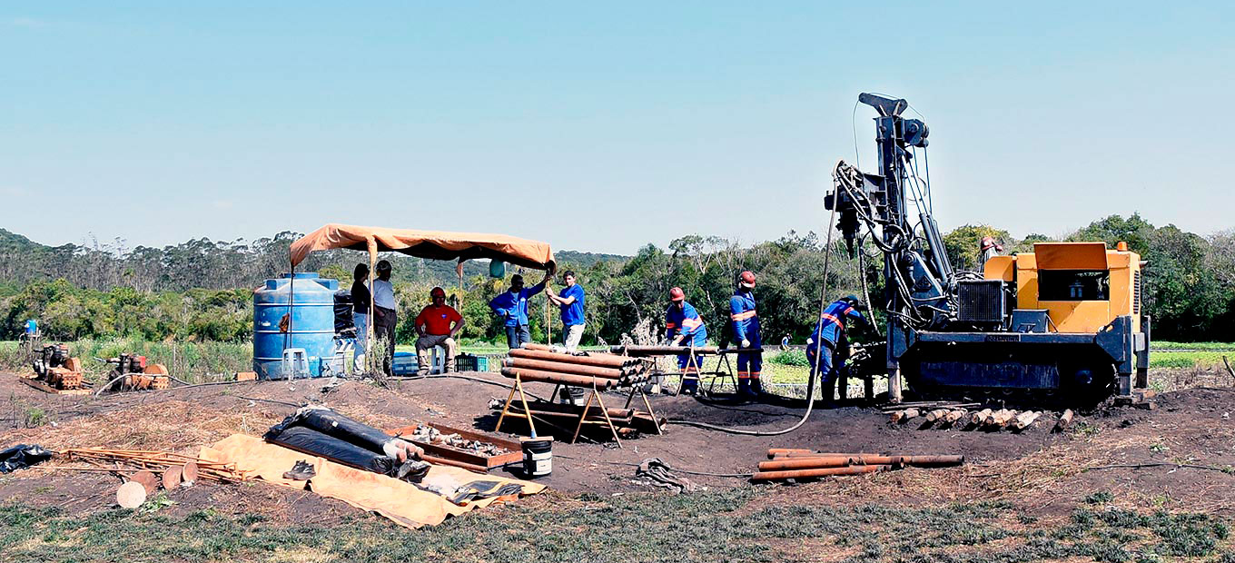 Em campo aberto equipamentos de perfuração com trabalhadores ao lado vestindo roupa azul e capacetes laranja, cavaletes com tubos empilhados e uma tenda com lona laranja e algumas pessoas sob ela, ao lado caixa de água azul, ao fundo vegetação.