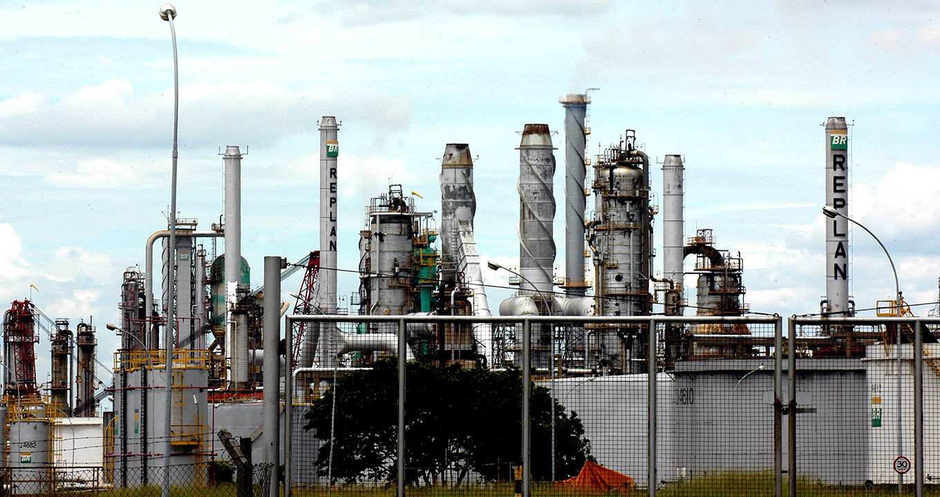 Em área externa, imagem panorâmica e frontal de vários tipos de torre, de diferentes alturas e diâmetros, que integram uma indústria pesada de refino de petróleo. Elas são de metal, a maioria em tom cinza, sendo que em duas delas está escrito BR Replan. Ao fundo, aparece o céu nublado, e à frente, parte de uma grande que faz a demarcação da área interna da empresa. Imagem 1 de 1.