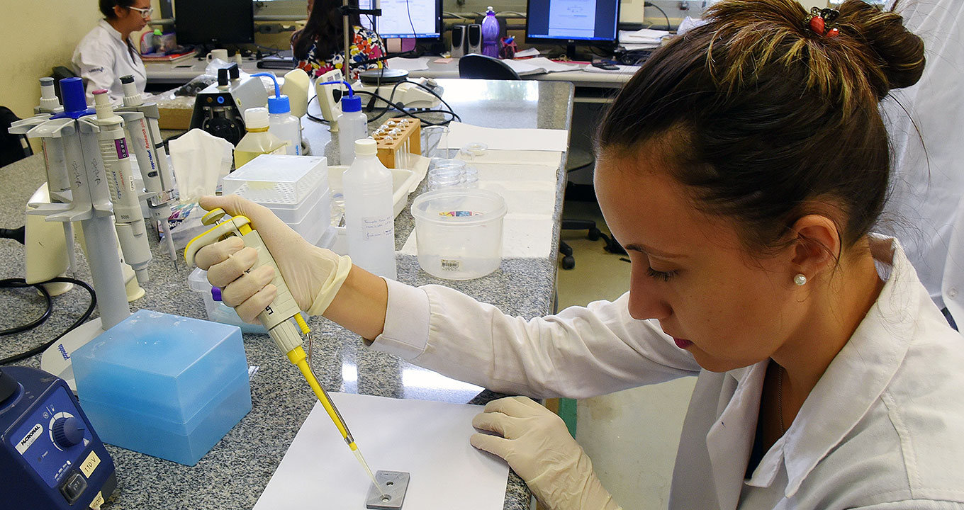 Audiodescrição: Em imagem close-up, em perfil, de cima para baixo, mulher em laboratório manipula biossensor segurando-o com a mão direita. À frente dela, há uma bancada retangular de granito cinza, sobre a qual encontram-se vários recipiente plásticos nos formatos quadrado e retangular. Ele veste jaleco branco. Imagem 1 de 1.