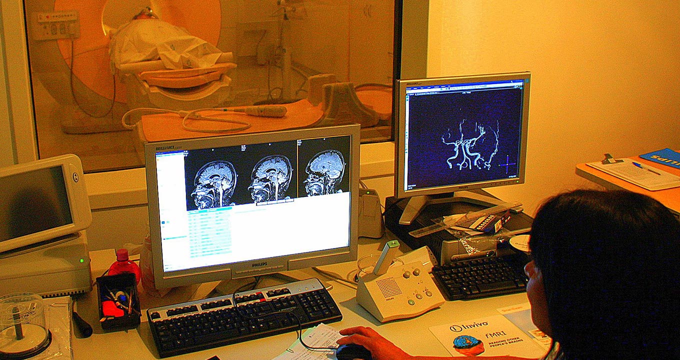 Audiodescrição: Imagem frontal, em sala de realização de exames por imagem, com pouca luz, mulher, à direita e de costas na imagem, observa dois monitores de lcd que estão sobre uma mesa retangular, sendo um ao centro e outro à direita. Um, ao centros, mostra três imagem de exame da região da cabeça e cérebro; o da direita uma imagem de cérebro. À frente dos monitores, ha uma parede com uma janela de vidro transparente com cerca de um metro por um metro, que permite  visualizar uma outra sala. Imagem 1 de 1