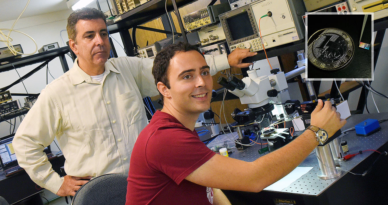 Em laboratório de física, imagem frontal e plano médio, dois homens, um ao lado do outro, sendo um em pé, à esquerda, e outro sentado, ao centro. À frente deles há uma bancada retangular de inox com vários equipamentos, onde se destaca um moderno microscópio, sendo que o homem ao centro manipula o equipamento com a mão direita, sorrindo. Ele veste camisa de mangas curtas em tom grená, enquanto o outro homem mantém a mão esquerda na cintura e veste camisa social de mangas compridas em tom bege. Imagem 1 de 1