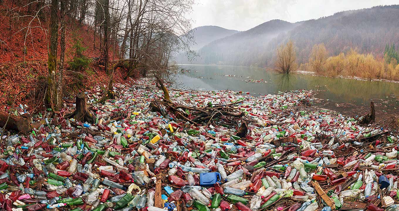 Audiodescrição: Em área externa, imagem panorâmica, às margens de um amplo rio, à direita, milhares de garrafas pet coloridas amassadas e acumuladas. À esquerda da imagem, há um barranco e algumas árvores, enquanto que ao fundo há uma densa floresta com árvores comuns da Europa e América do Norte. O tempo está parcialmente nublado. Imagem 1 de 1.